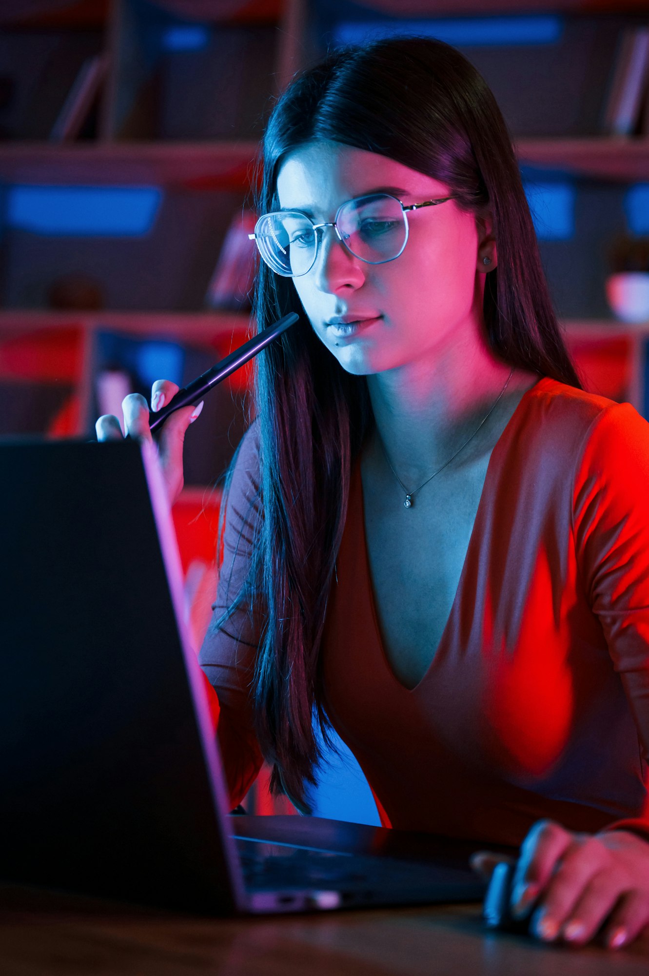 Beautiful woman in glasses and red wear is sitting by the laptop in dark room with neon lighting